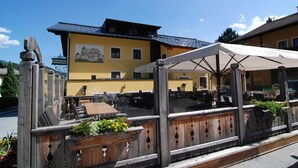 Een terras met houten tafels en stoelen, een houten hek en een gebouw met een gele gevel op de achtergrond.