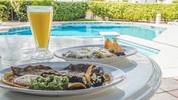 Mesa de jantar com duas porções de comida e um copo de suco de laranja perto de uma piscina.