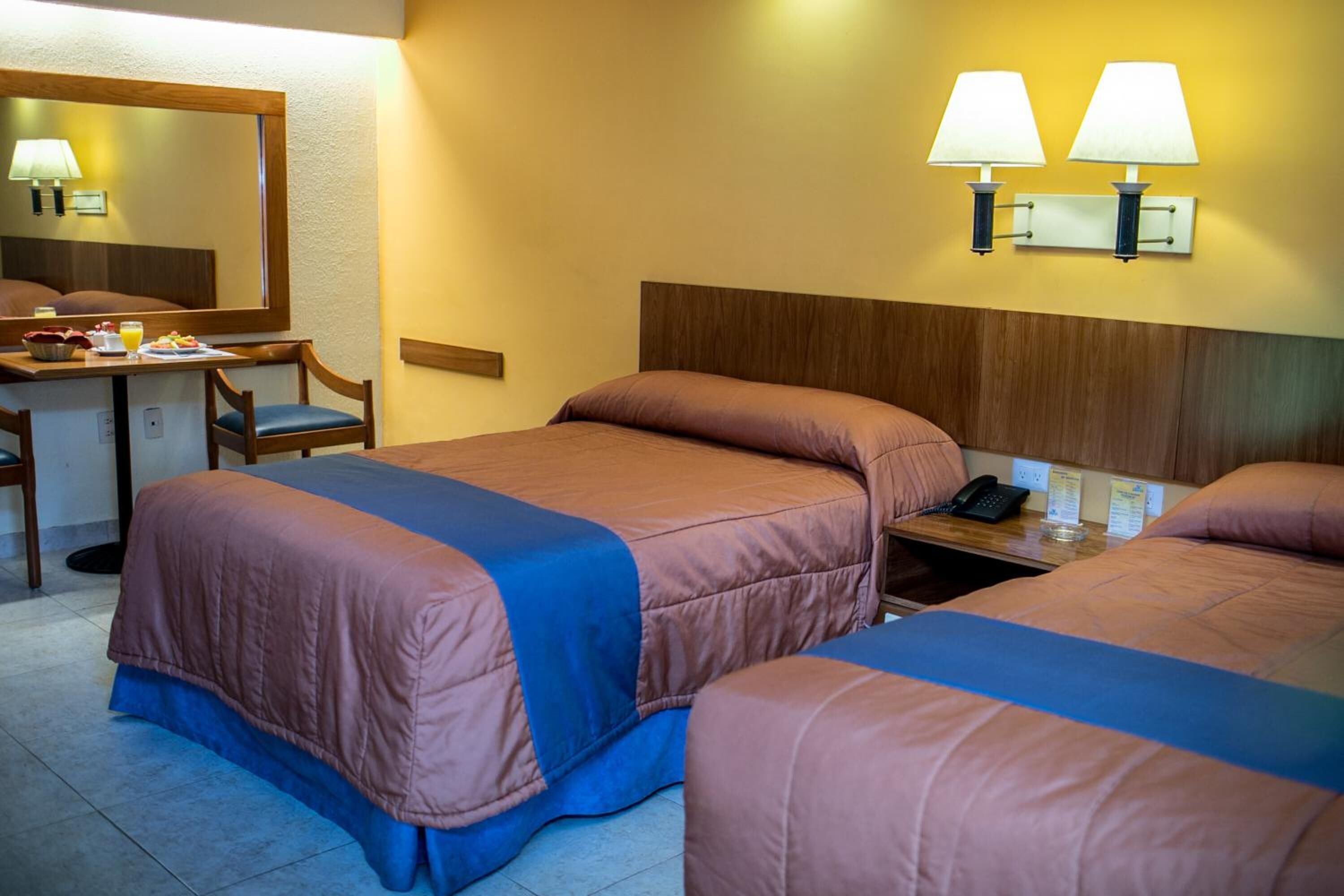 Habitación de hotel con dos camas, un escritorio con una silla, un espejo y dos lámparas de pared.