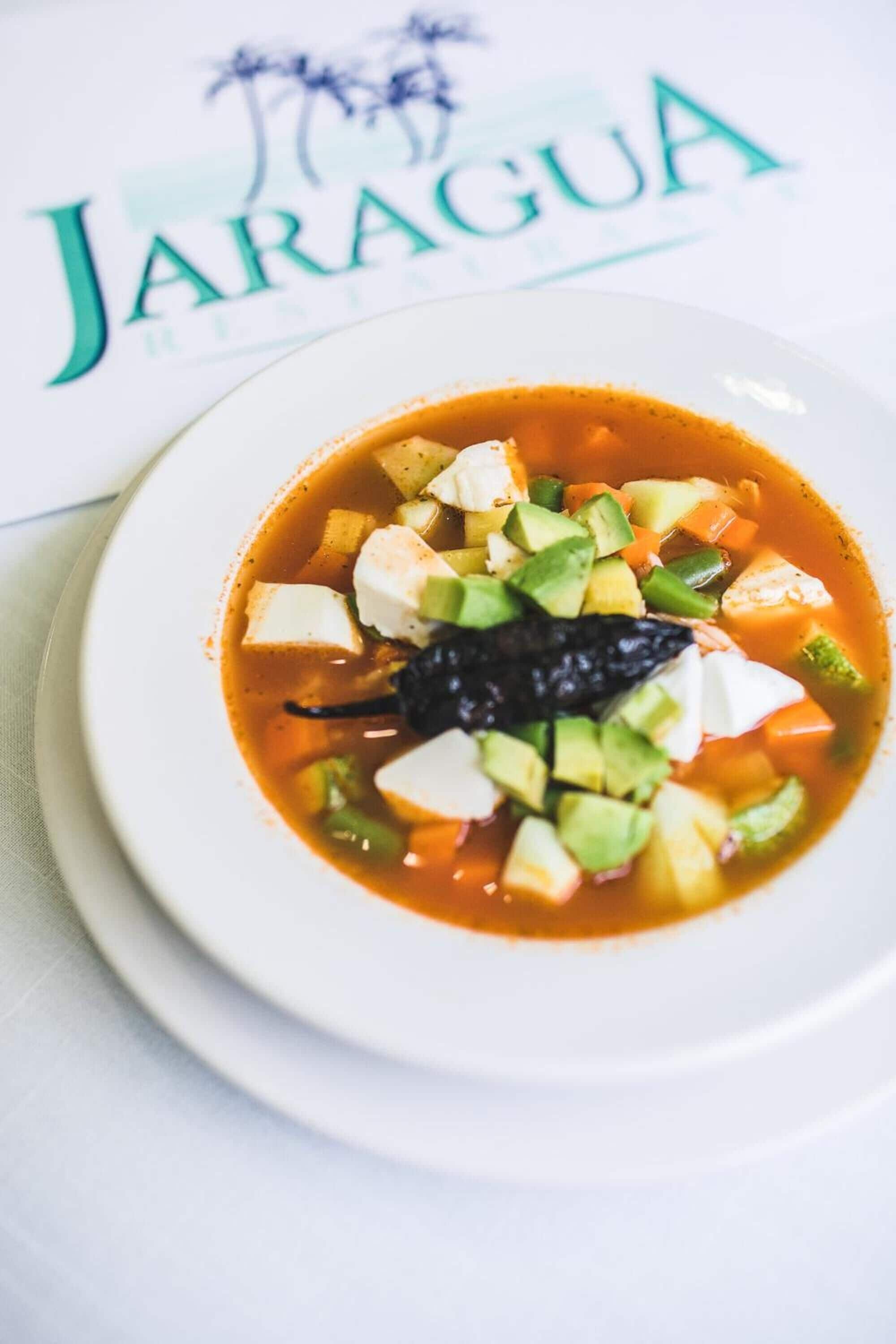 A bowl of soup with avocado and a black pepper on a white plate, with a menu in the background.