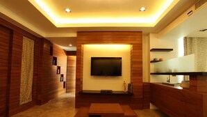 Een moderne woonkamer met een flatscreen televisie aan een houten wand, een houten salontafel en een inbouwkeuken met een magnetron en een klein plankje.