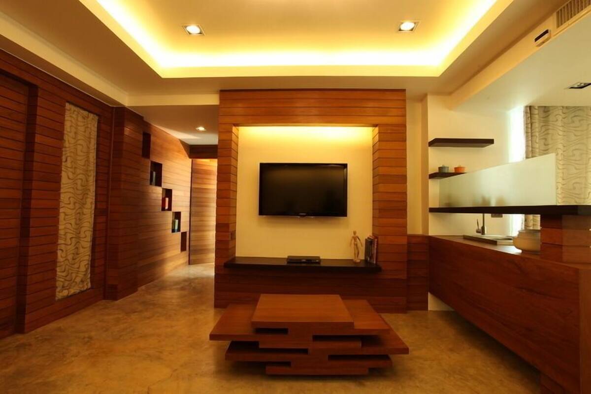 Una sala de estar moderna con un televisor de pantalla plana montado en una pared de madera, una mesa de centro de madera y una encimera de cocina integrada con un microondas y un estante pequeño.