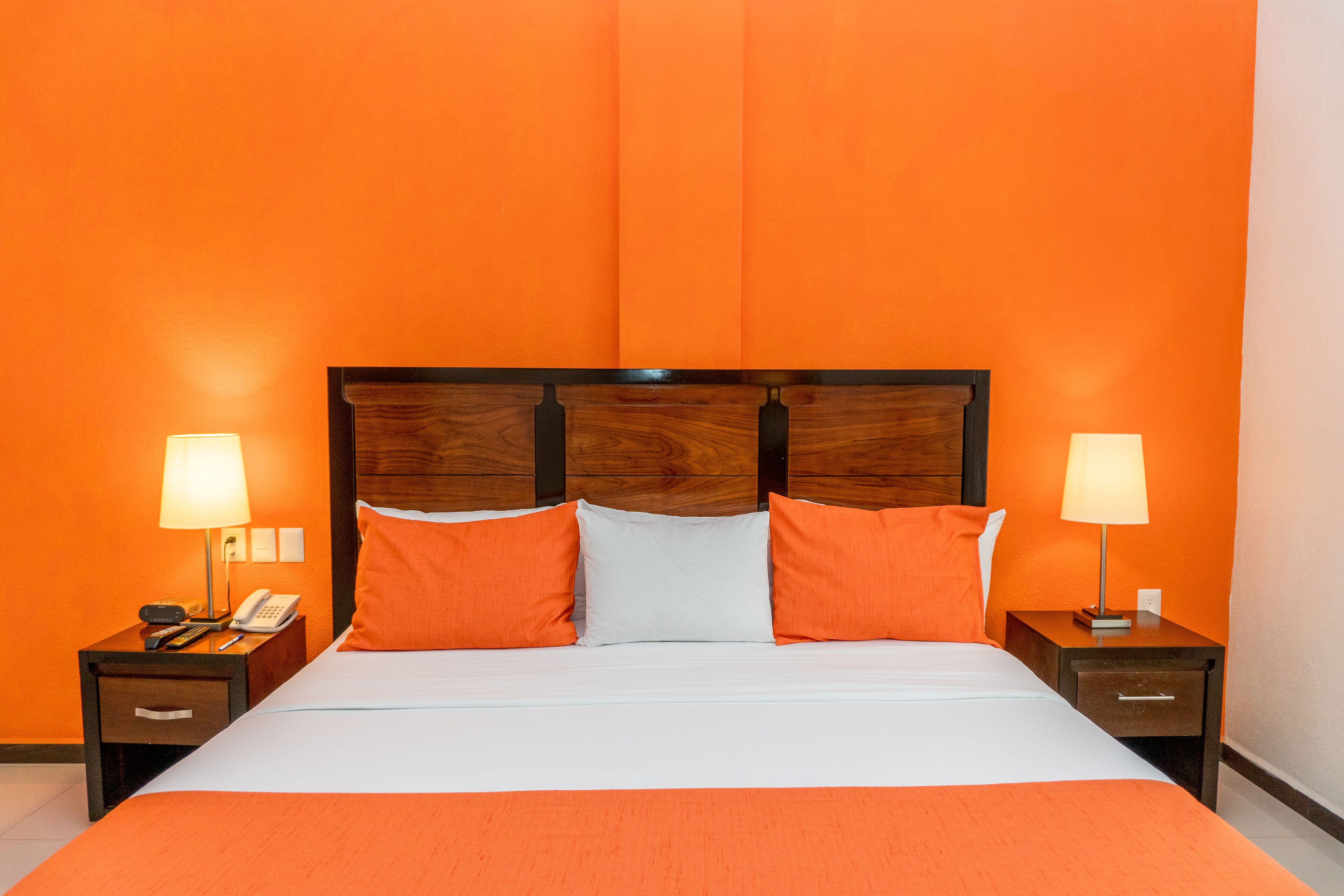 Habitación de hotel con pared naranja, cabecera de madera, cama con ropa de cama blanca y naranja, dos mesitas de noche con lámparas y un teléfono sobre una de ellas.
