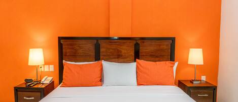 Habitación de hotel con pared naranja, cabecera de madera, cama con ropa de cama blanca y naranja, dos mesitas de noche con lámparas y un teléfono sobre una de ellas.