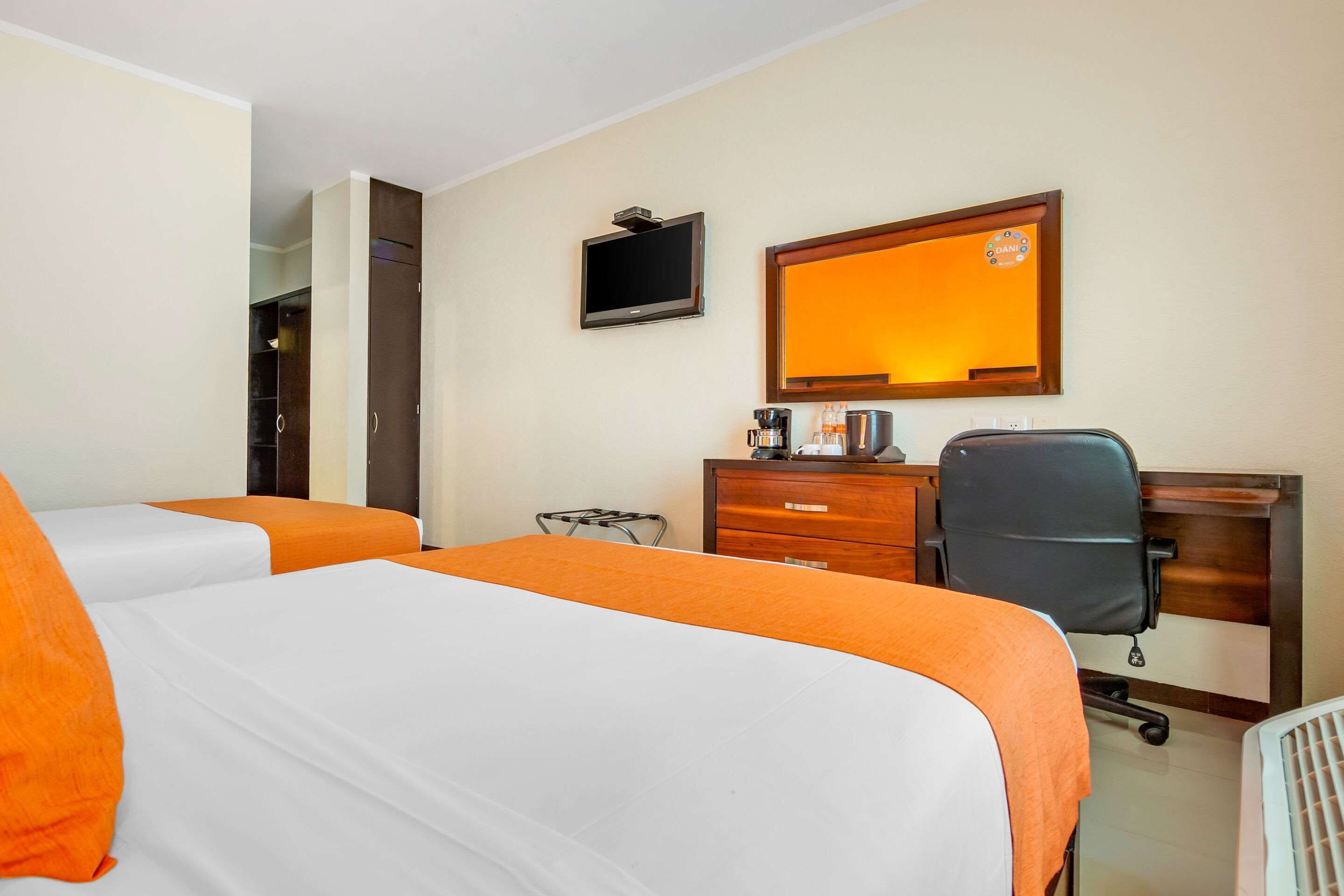 Quarto de hotel com duas camas, uma escrivaninha, uma cadeira, uma televisão e um espelho.