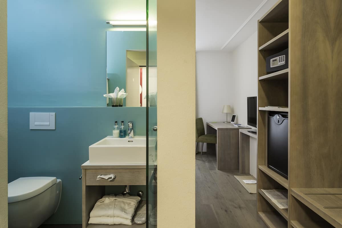 A modern bathroom with a toilet, sink, and mirror. There is a wooden shelf with a television and a desk with a lamp.