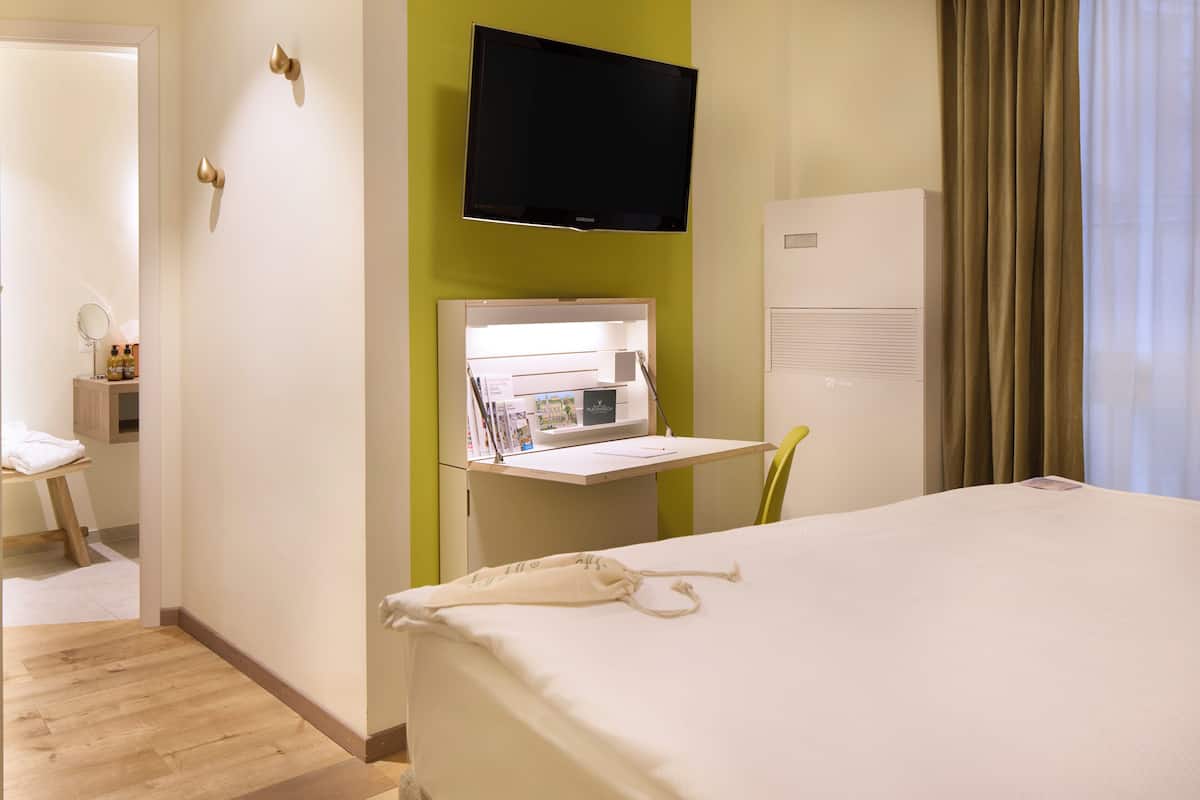 A hotel room with a bed, a desk with a flat-screen TV, a minibar, and a window with curtains.
