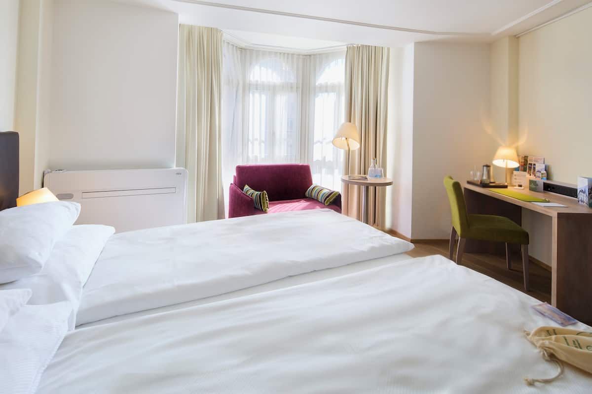 A hotel room with a large bed, a desk, a chair, a lamp, and a window with curtains.