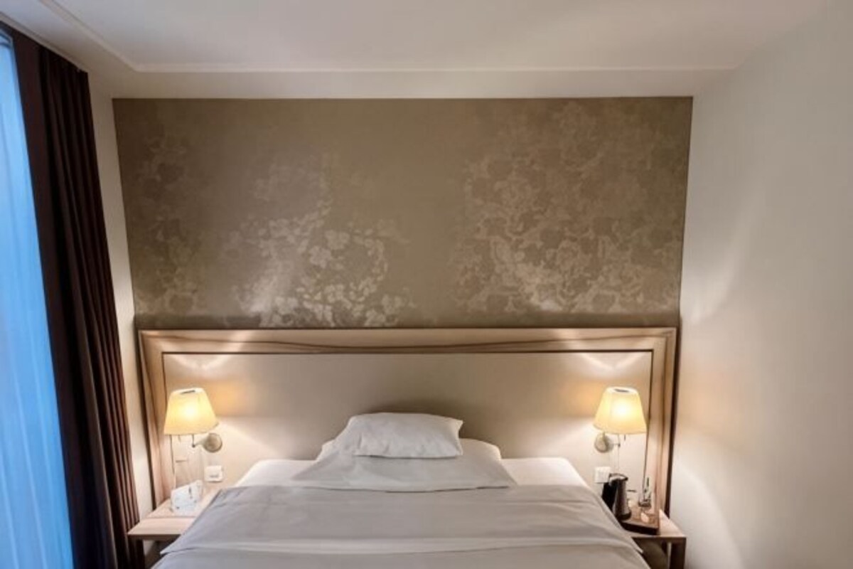 A hotel room with a large bed, two bedside lamps, a headboard, and a plain wall.