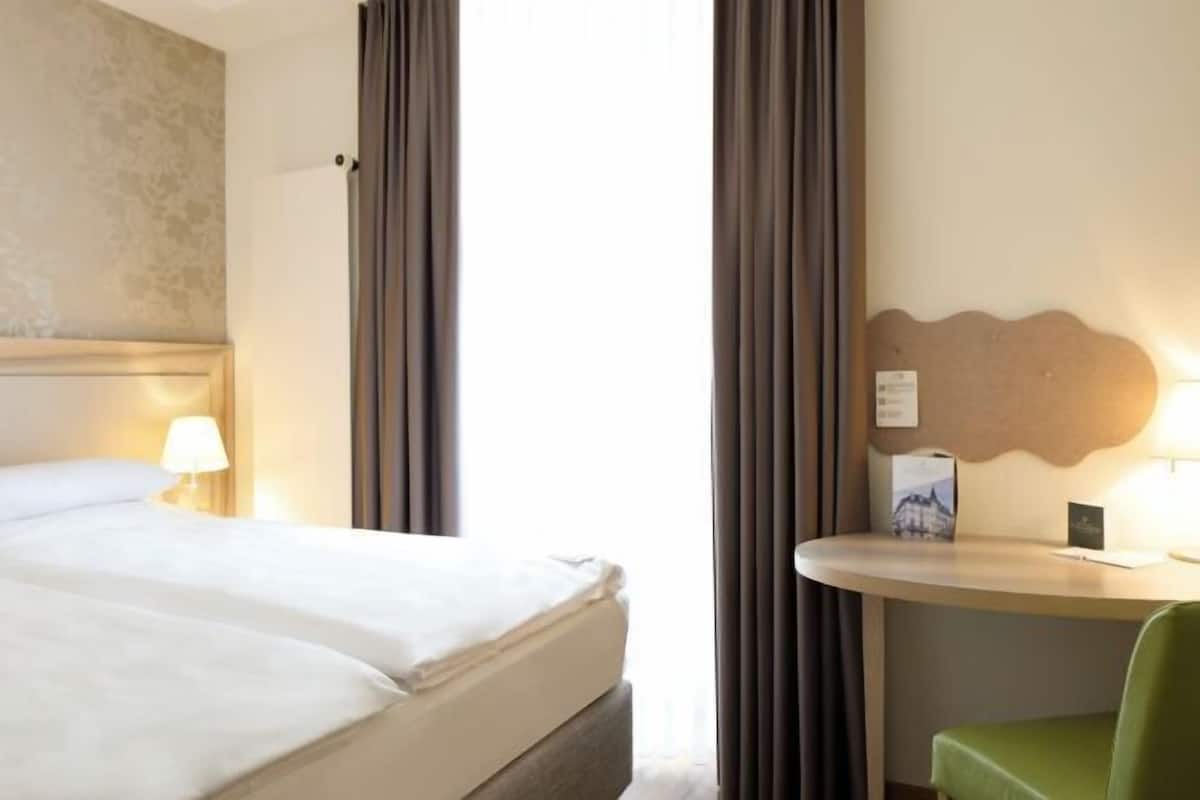 A hotel room with a bed, a desk with a lamp, curtains, and a wall-mounted light.