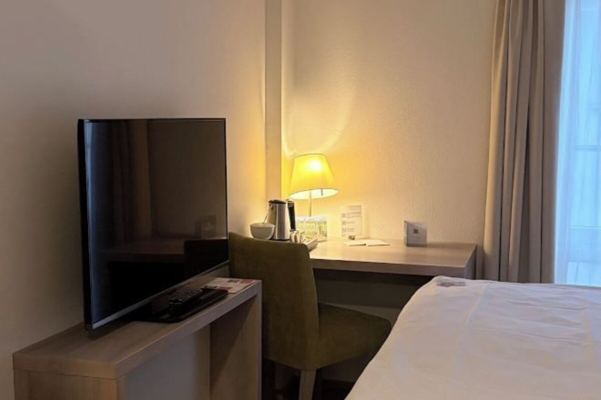 A hotel room with a bed, a desk with a lamp, a television, and a chair.