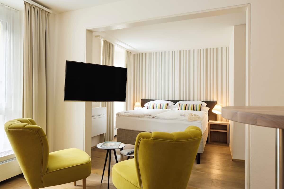 A hotel room with a bed, a TV mounted on the wall, and two yellow chairs.