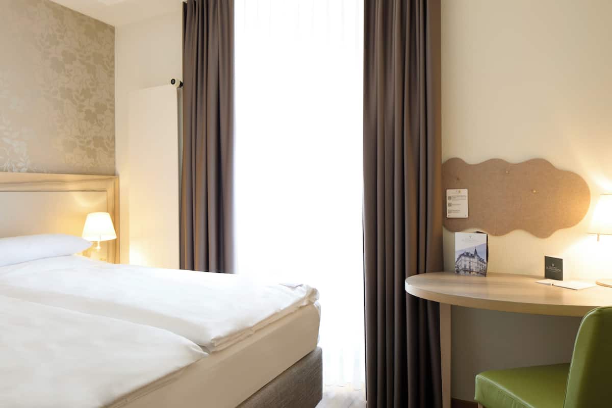 A hotel room with a bed, a desk, a chair, and a wall-mounted lamp.