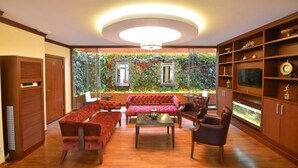 Un salon moderne avec un canapé rouge, une table basse en verre et des fauteuils en cuir. Une grande fenêtre offre une vue sur un mur végétal et des plantes.