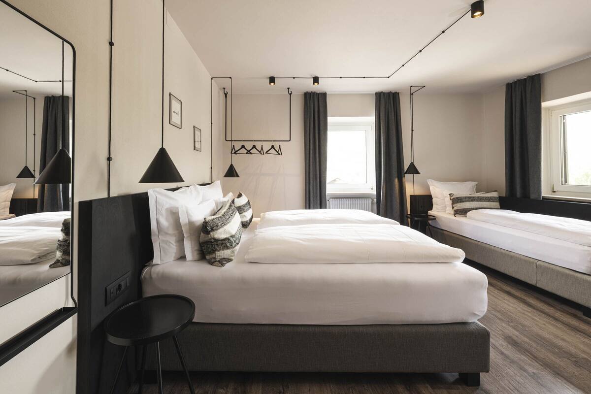 A modern hotel room with a large bed, a smaller bed, a desk, and a chair. There is a window with curtains, a wall-mounted light fixture, and a framed picture on the wall.