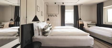 A modern hotel room with a large bed, a smaller bed, a desk, and a chair. There is a window with curtains, a wall-mounted light fixture, and a framed picture on the wall.
