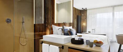 A modern hotel bathroom with a glass-enclosed shower, a large mirror, a vanity with a sink, and a bedside table with toiletries.