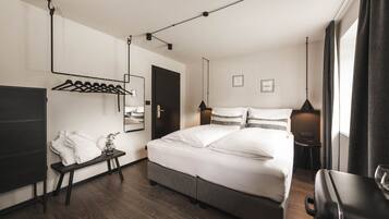 Quarto de hotel moderno com uma cama grande, mesa de cabeceira com luminária, cabideiro e espelho.