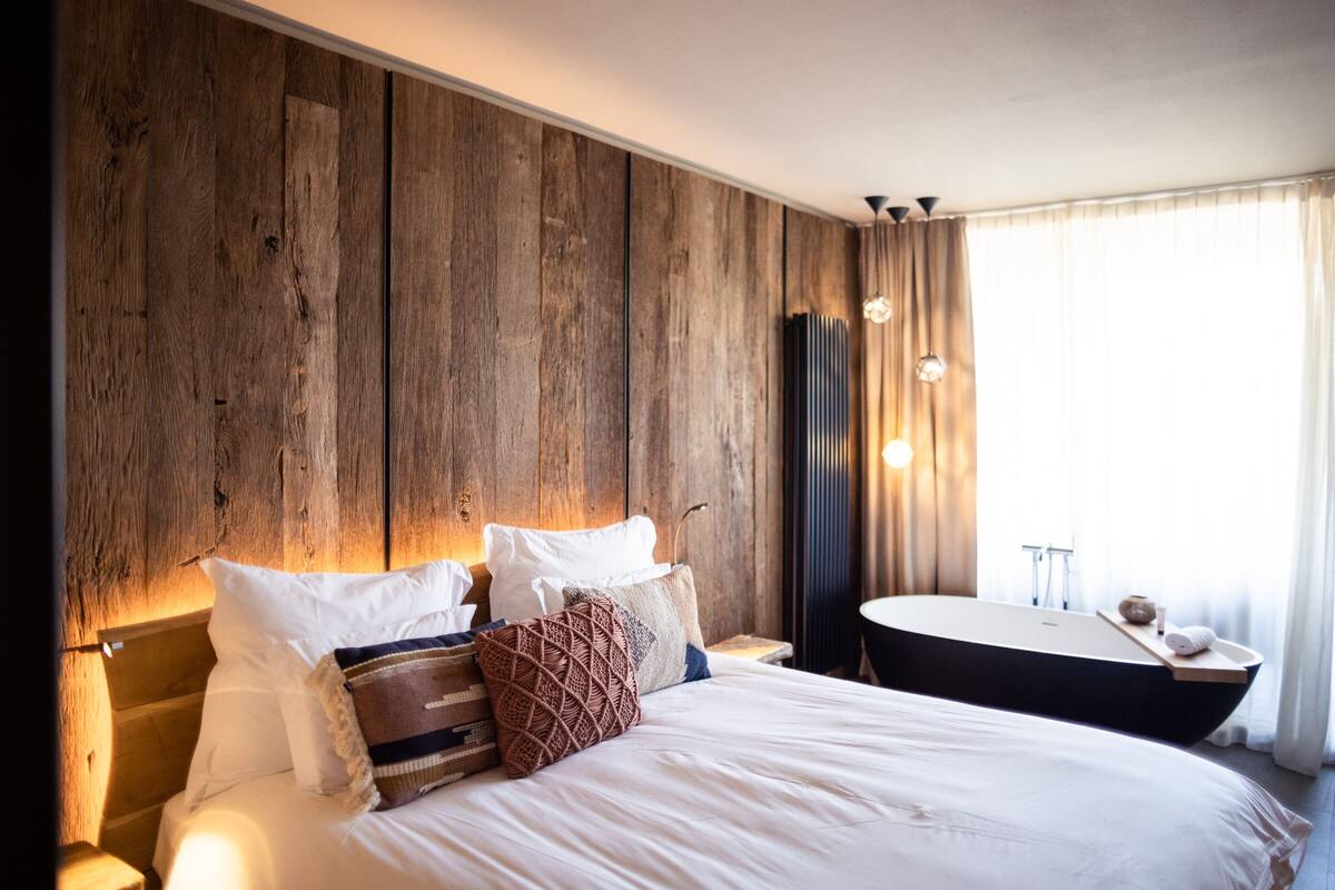 A large bed with white linens, a wooden headboard, and multiple pillows. There is a round mirror and a small table with a vase and a towel.