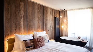 A large bed with white linens, a wooden headboard, and multiple pillows. There is a round mirror and a small table with a vase and a towel.