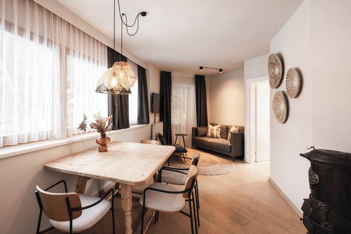 A dining area with a wooden table and chairs, a hanging light fixture, and a vase with dried flowers.