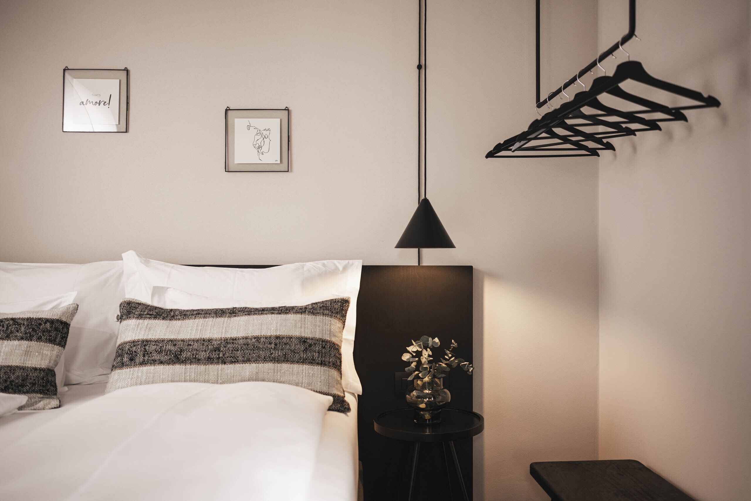 A neatly made bed with striped pillows, a small black side table with a vase, and a black hanger rack against the wall.