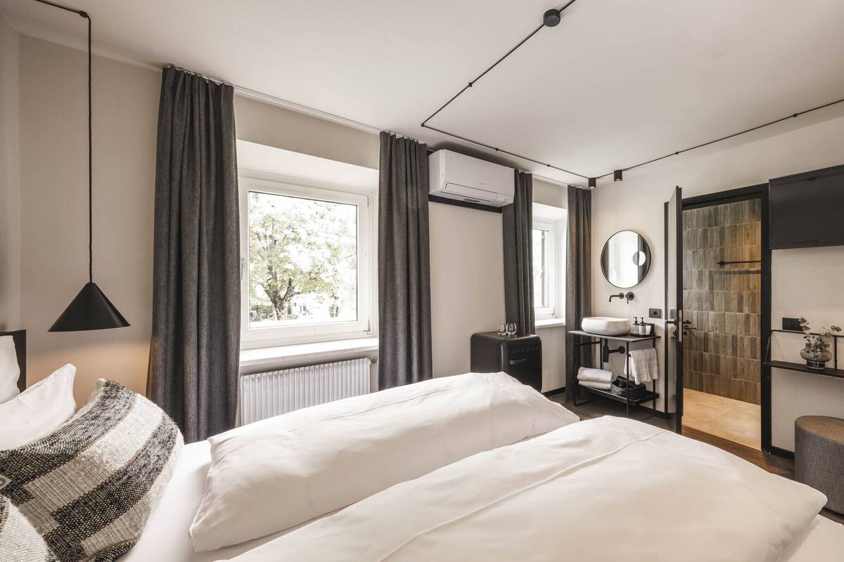 A modern hotel room with a large bed, a bedside table, a mirror, a window with curtains, and a view of trees outside.