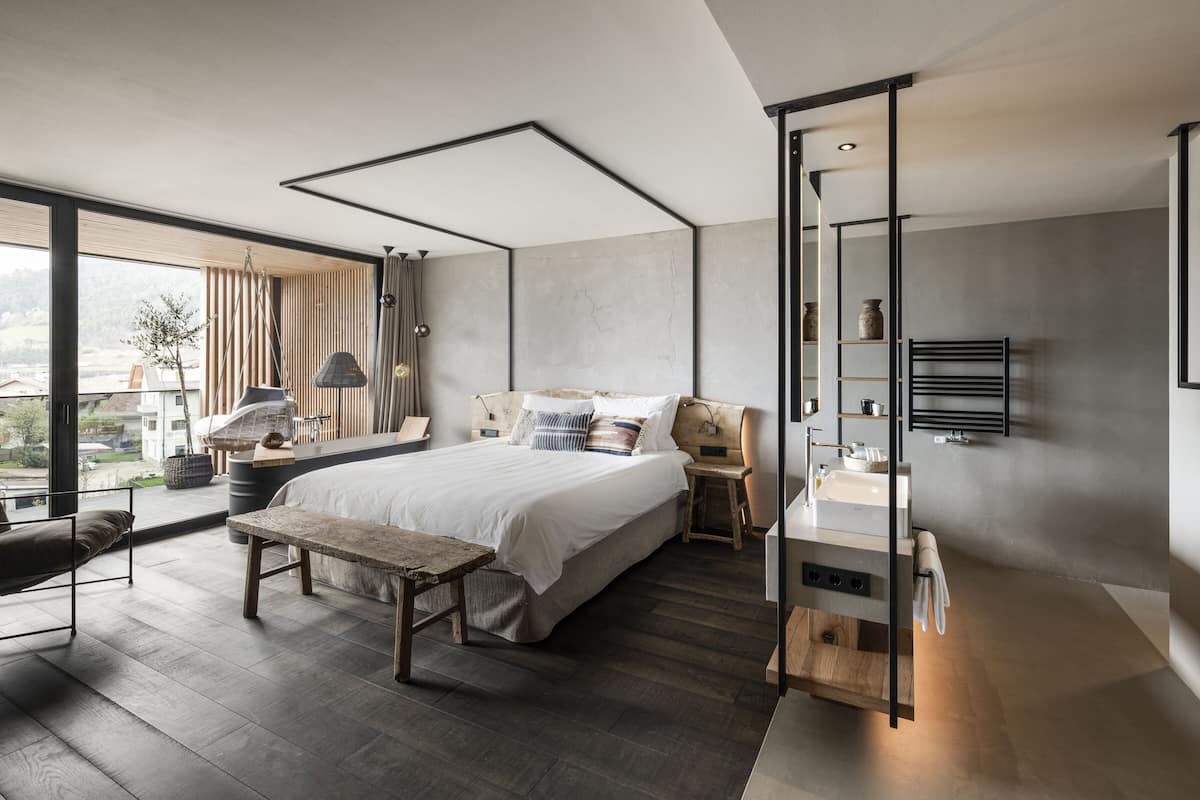 A modern bedroom with a large bed, a wooden bench, a desk, and a bathroom with a towel rack.