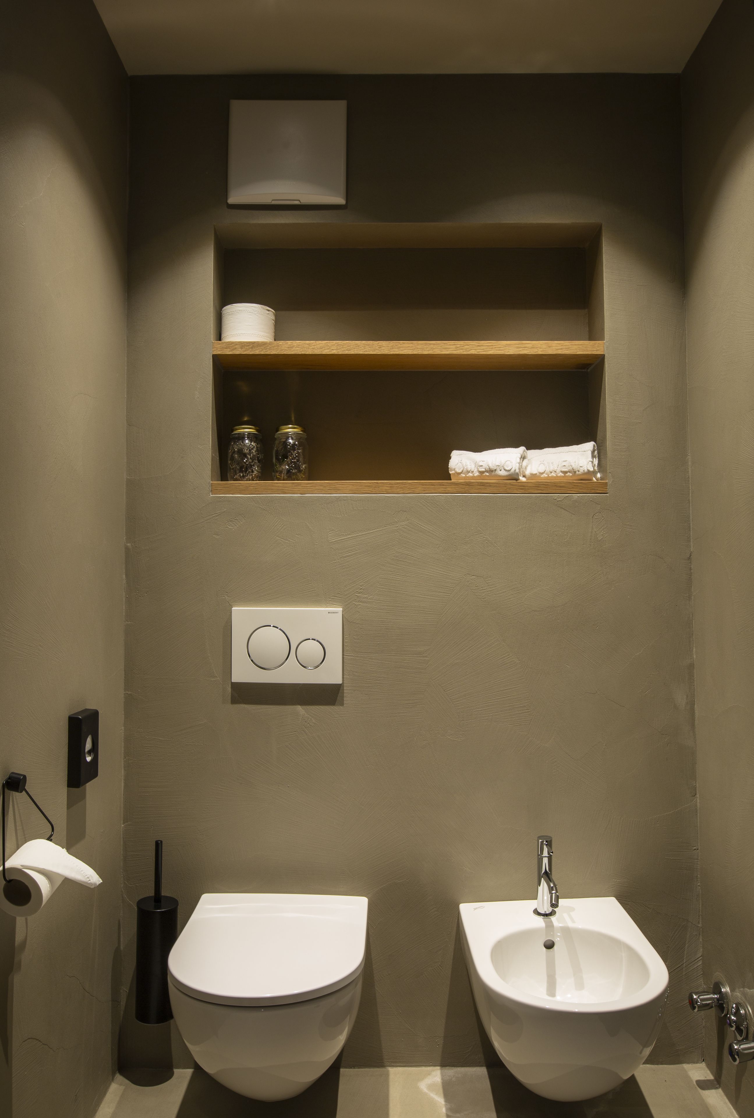 Un baño con dos lavamanos, un inodoro, una repisa con artículos de aseo y una luz empotrada en la pared.