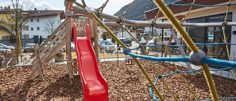 A playground with a red slide, surrounded by wooden play structures and a rope net.