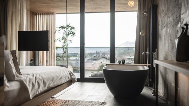 A modern bedroom with a large bed, a freestanding bathtub, and a city view through floor-to-ceiling windows. - Löwenhof (Varna)