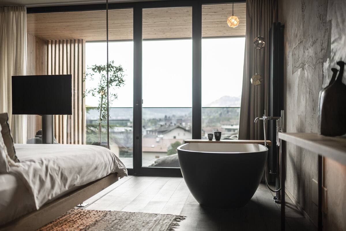 A modern bedroom with a large bed, a freestanding bathtub, and a city view through floor-to-ceiling windows.