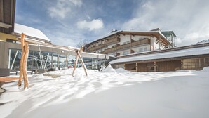 Área externa coberta de neve com baloiço de madeira, um edifício de vidro e um prédio de vários andares com varandas.