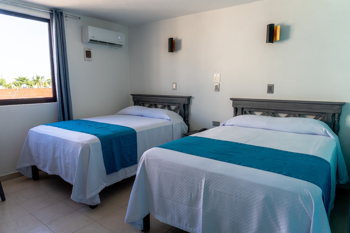 A hotel room with two single beds, a window with curtains, an air conditioning unit, and a wall-mounted light fixture.