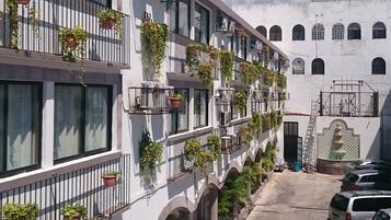 Un edificio de varios pisos con balcones decorados con plantas en macetas, un estacionamiento con varios coches, y un cielo despejado con nubes.
