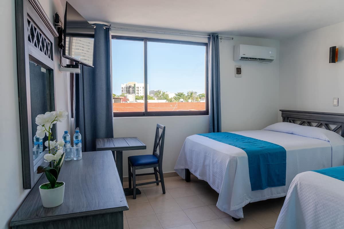 Hotelkamer met twee bedden, een bureau, een stoel, een potplant en een groot raam met uitzicht op gebouwen en palmbomen.