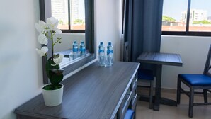 A hotel room with a wooden desk, a potted orchid, water bottles, a mirror, a window with curtains, and a view of buildings. - Hotel Hacienda de Castilla (Cancun)