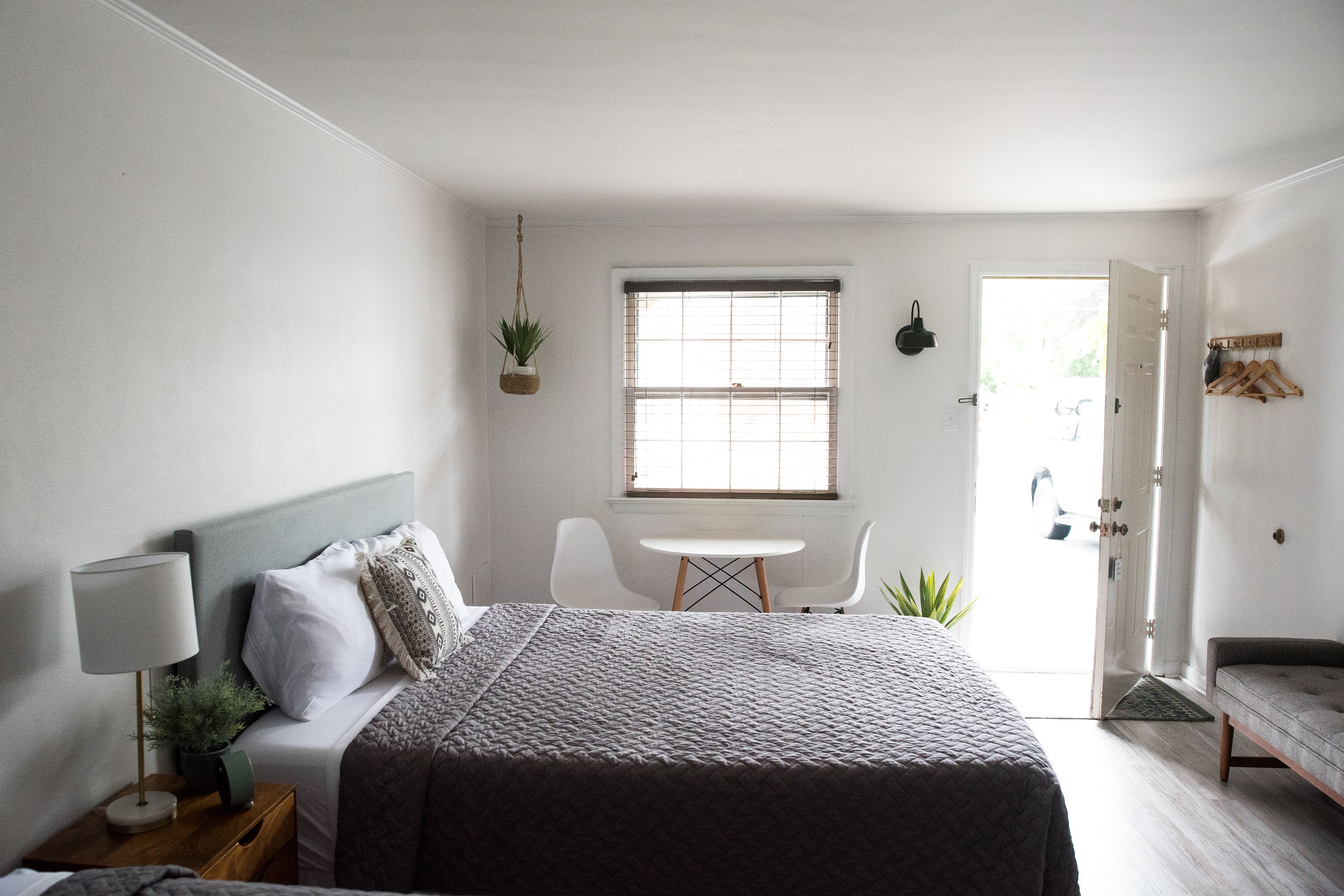 Quarto com cama, mesa de cabeceira, luminária, planta pendente, janela e uma pequena mesa com cadeiras.