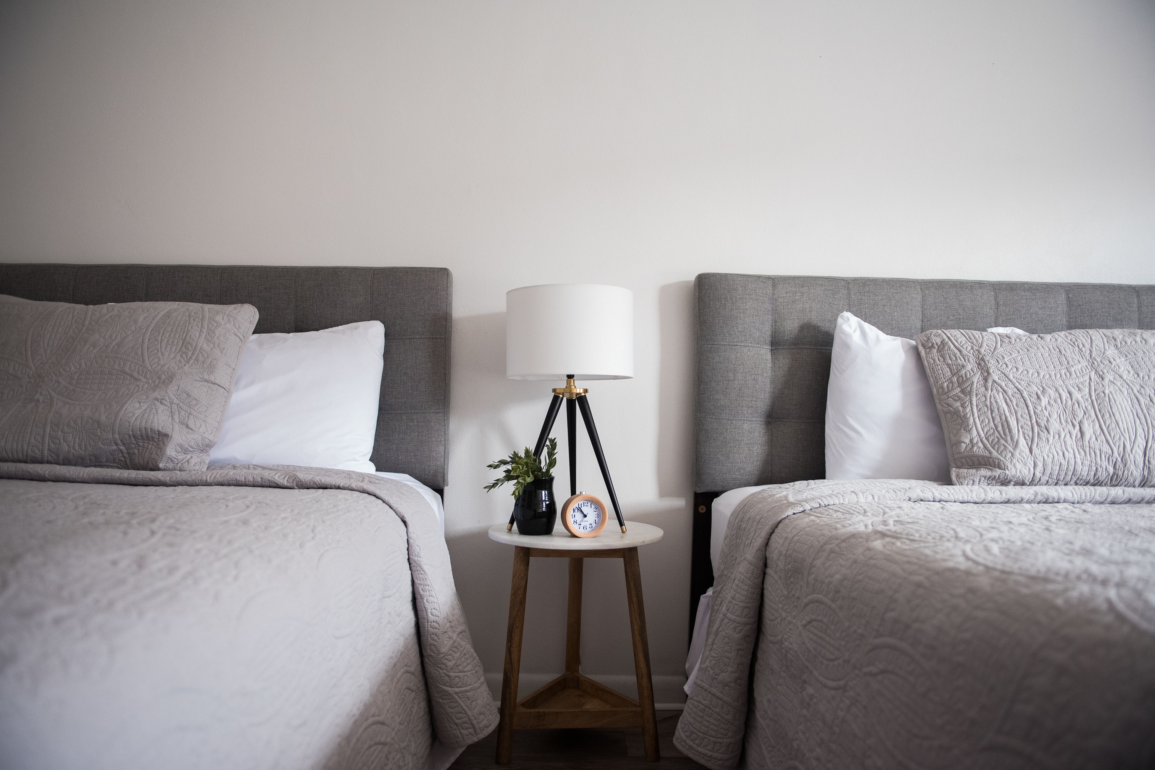 Ein Hotelzimmer mit zwei Betten, jeweils mit grauem Kopfteil und weißer Bettwäsche. Zwischen den Betten steht ein kleiner runder Tisch mit einer Lampe und einer Uhr.