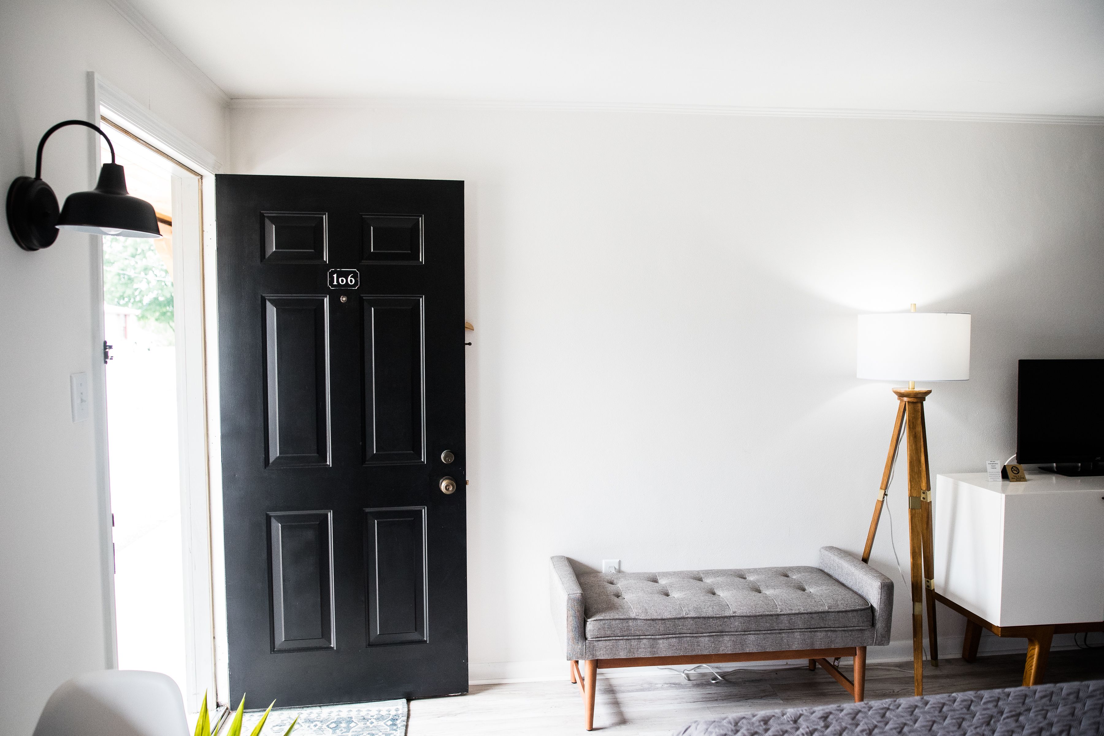 Quarto com porta preta, um banco estofado cinza, escrivaninha branca e abajur.