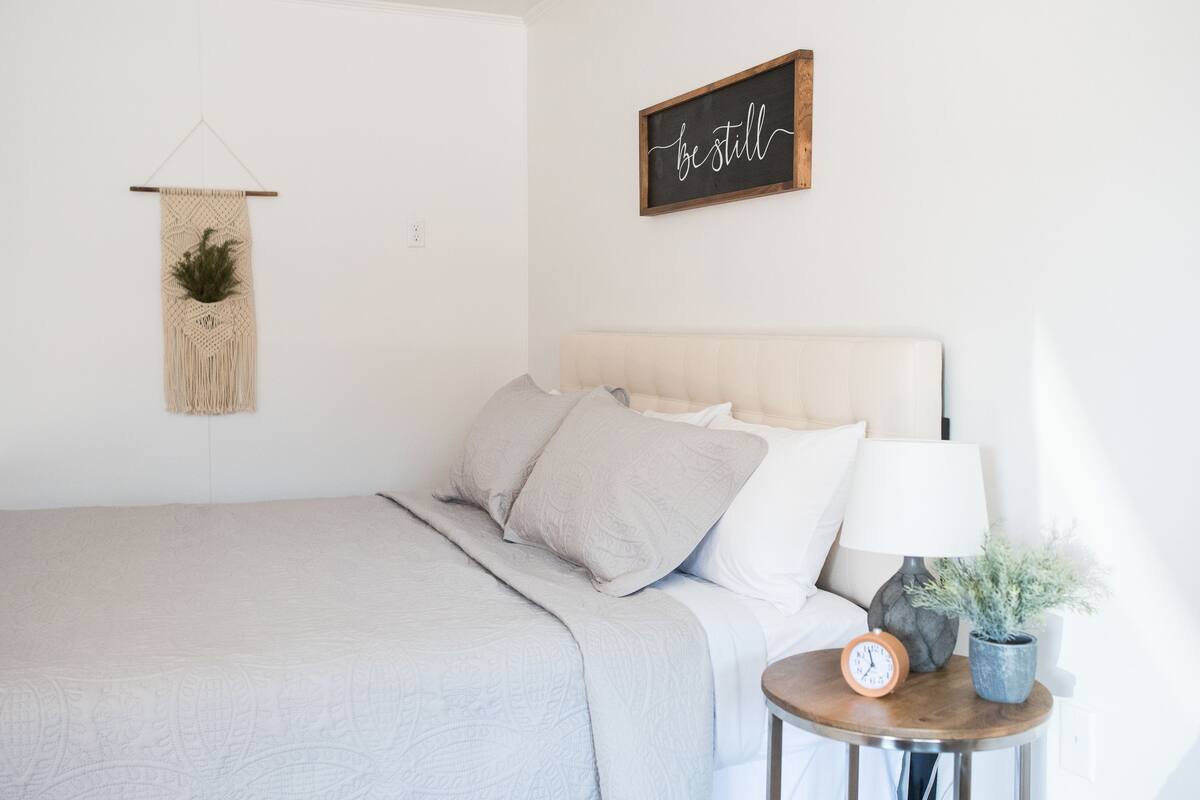 A neatly made bed with a headboard, a bedside table with a lamp and a clock, a macrame wall hanging, and a motivational sign on the wall.