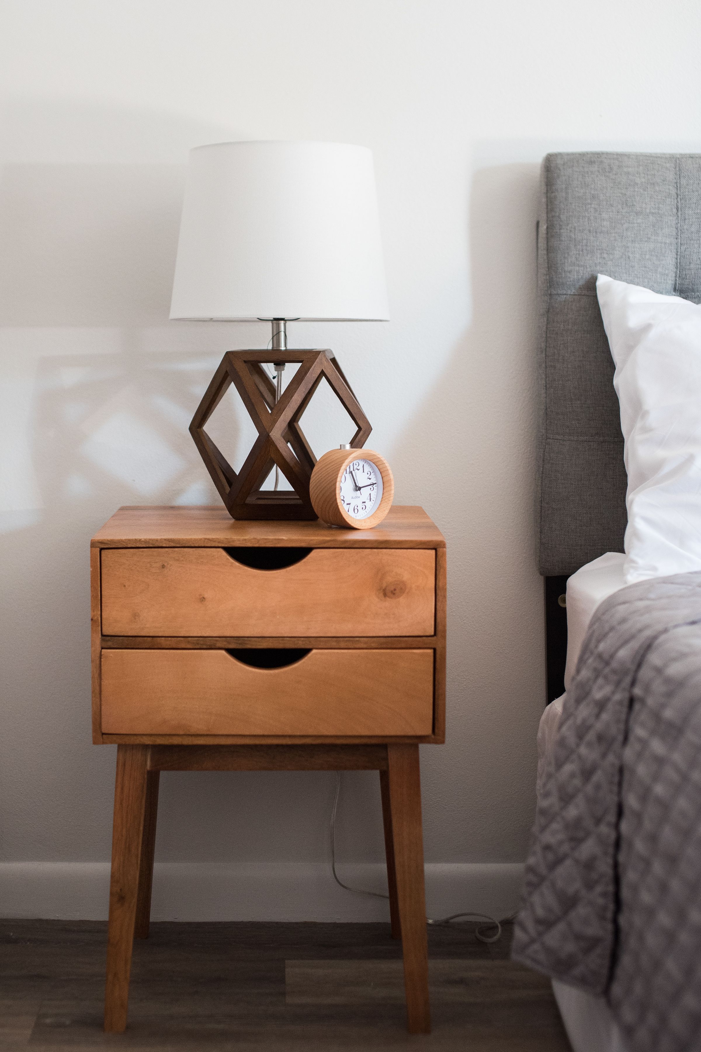 A wooden nightstand with a white lamp and a wooden clock.