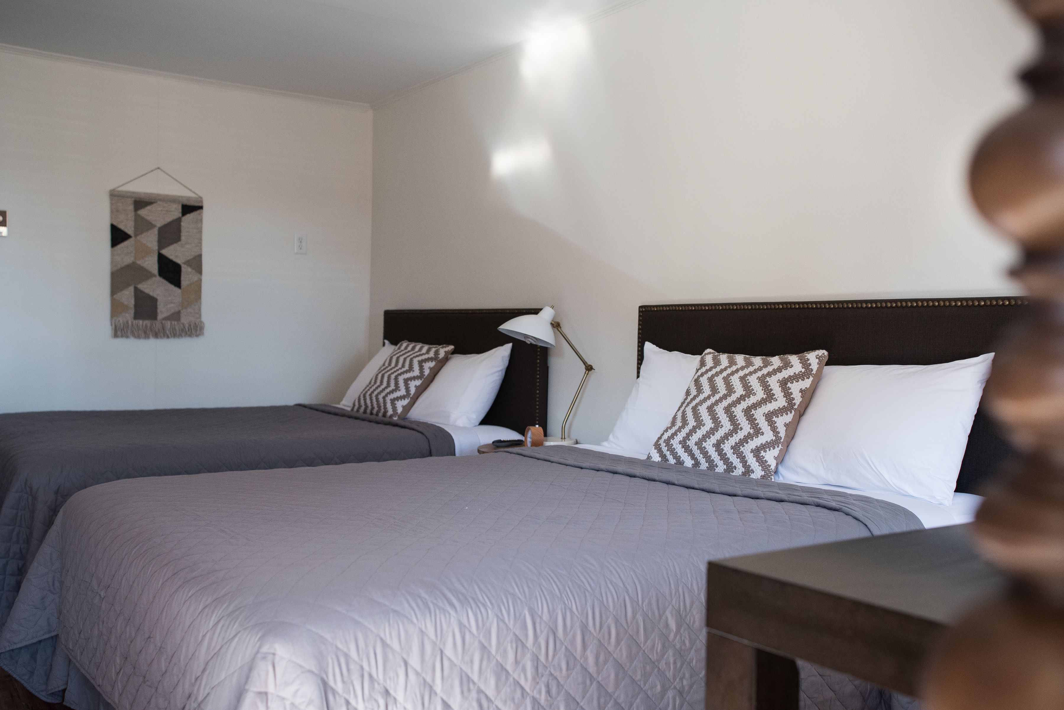 A hotel room with two beds, each with a headboard and pillows, a bedside table, and a wall-mounted lamp.
