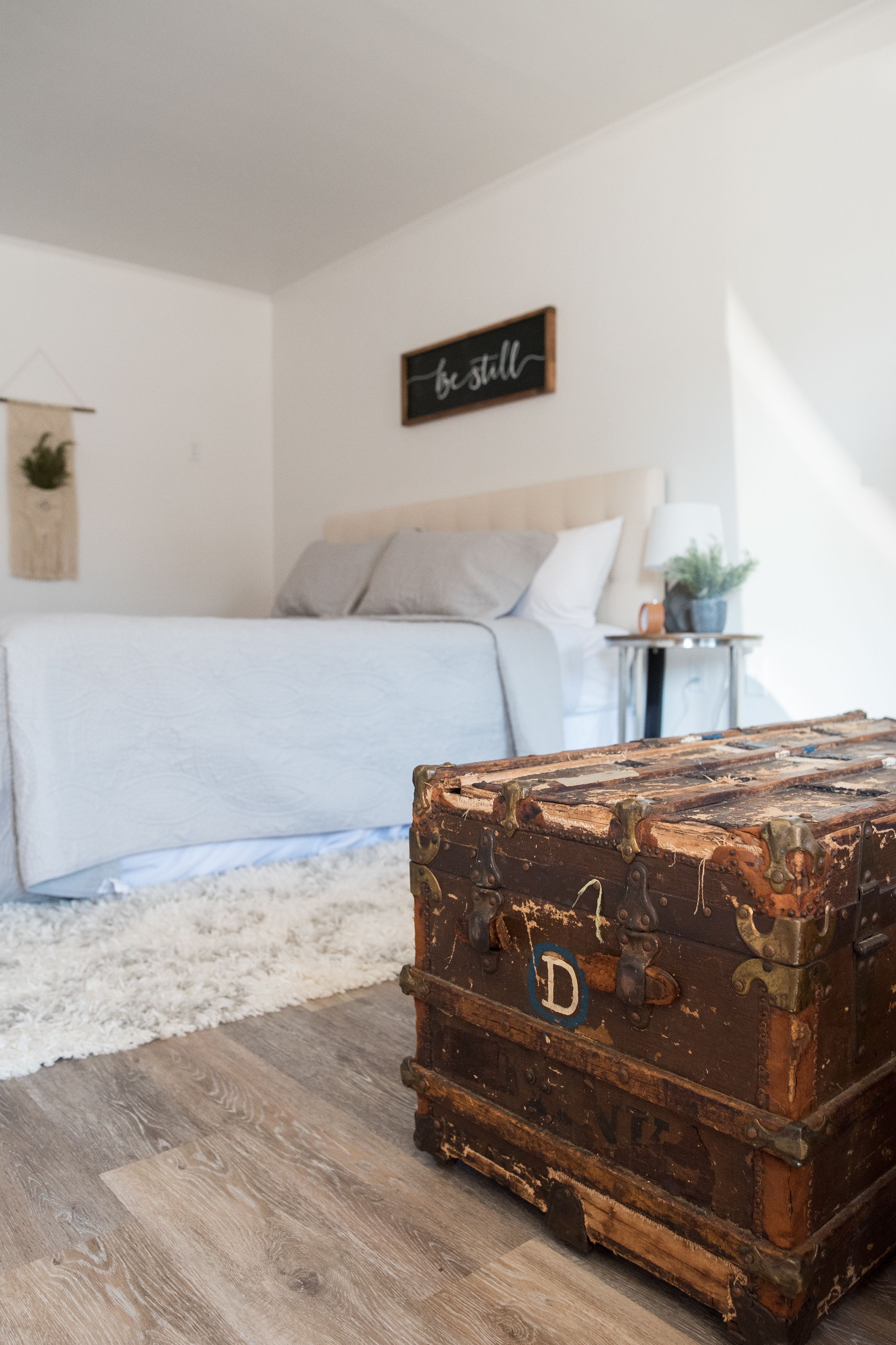 A bedroom with a large bed, a wooden floor, a vintage suitcase, a bedside table with a lamp, and a wall sign.