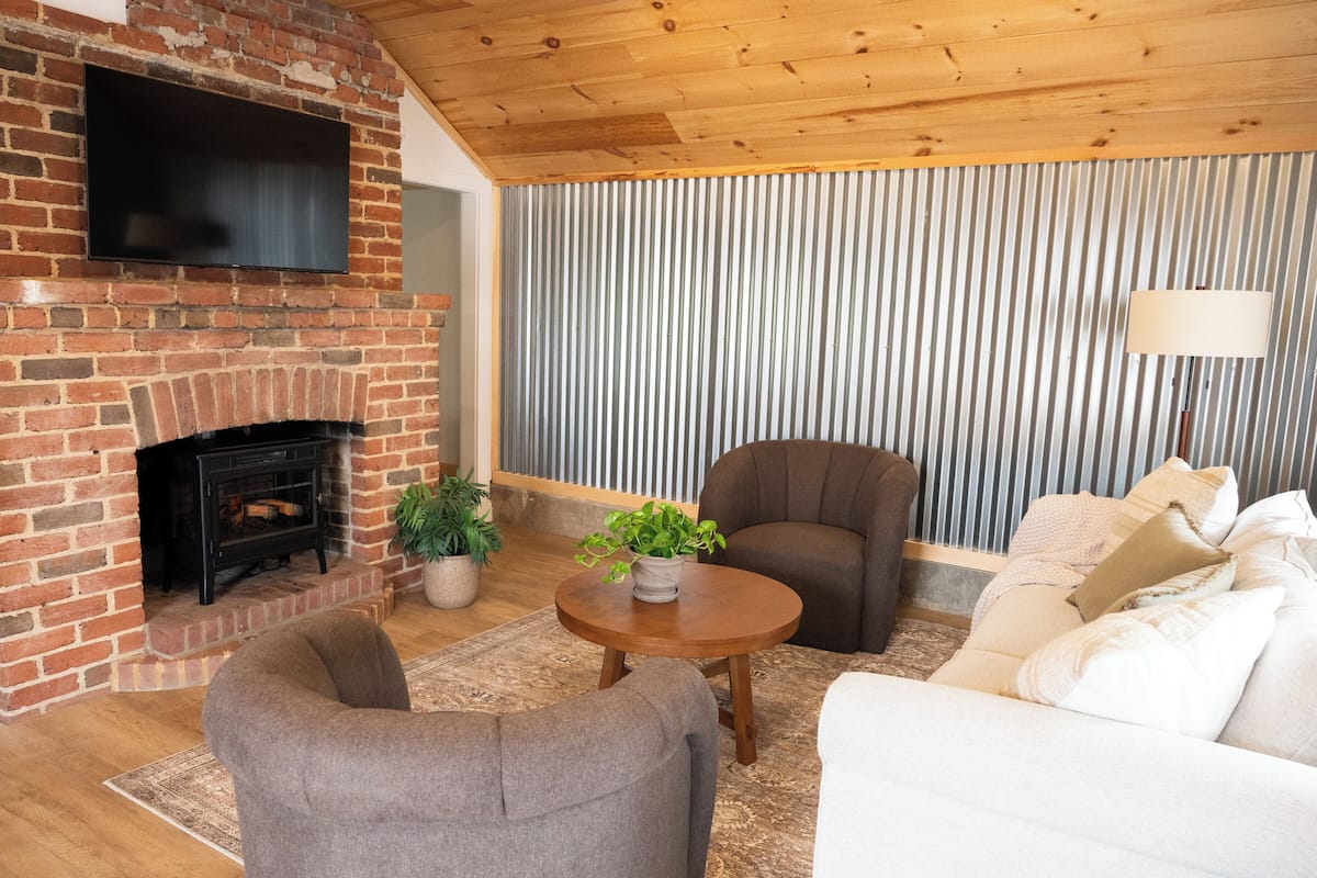 A living room with a brick fireplace, a flat-screen TV mounted above it, a round wooden coffee table, two armchairs, a white sofa, and a striped wallpaper backdrop.