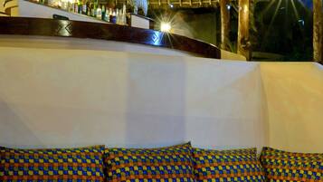 A bar area with a thatched roof, a sofa with patterned cushions, and a table with drinks and a candle.