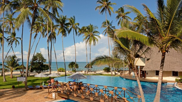 A pool area with a wooden bridge, palm trees, and a view of the ocean.