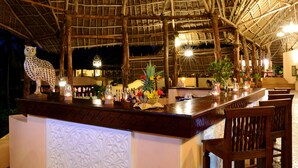 A bar area with a thatched roof, wooden bar counter, and seating. There are bottles and glasses on the counter, and a decorative owl statue.