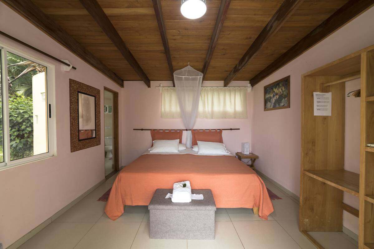 Ein Schlafzimmer mit einem großen Bett, einem Nachttisch, einem kleinen Regal und einem Fenster mit Vorhängen. Die Decke ist aus Holz.