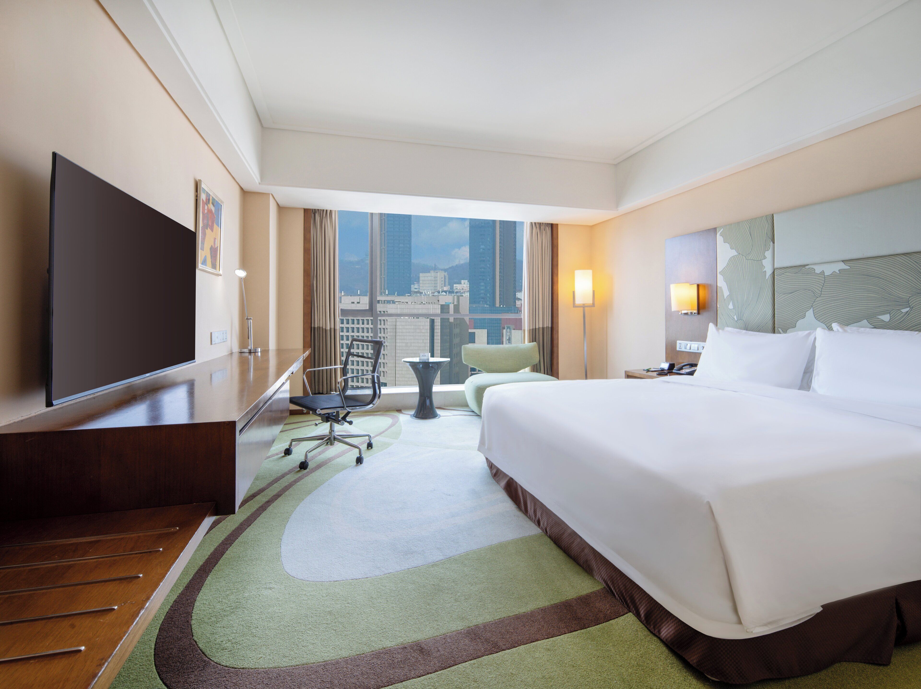 A hotel room with a large bed, a desk, a chair, a television, and a view of the cityscape.